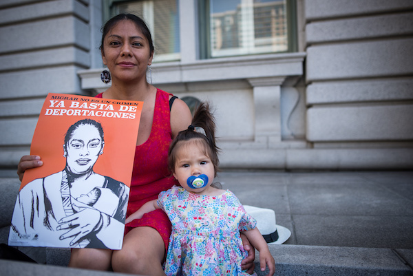 Mother and baby sitting on steps. Mother holds a sign that reads migrar no es un crimen. Ya basta de deportaciones.