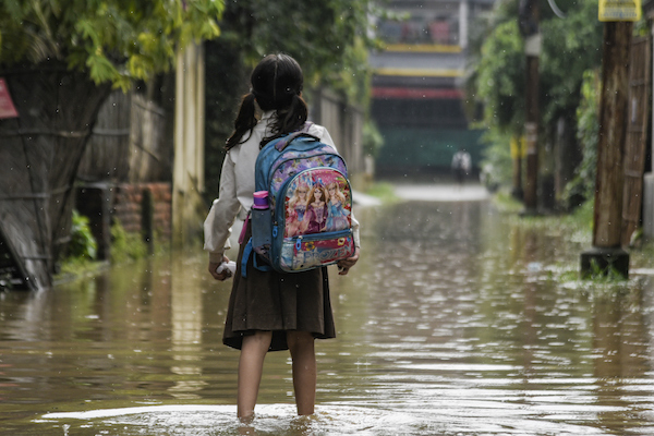 A child holding a backpack wading through a flooded street in India.